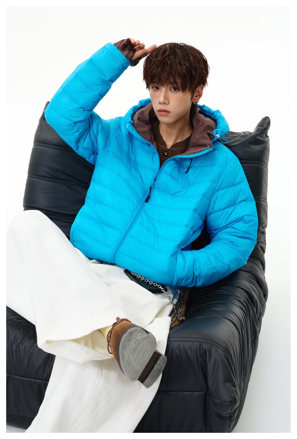 Model seated in a black chair wearing a bright cyan quilted hooded jacket with brown lining and white trousers.