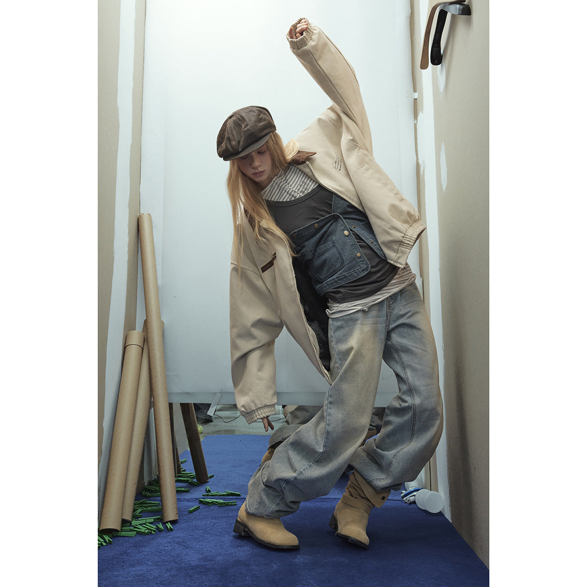 Model in a cream zip jacket with brown collar, layered tops, faded wide-leg jeans, and brown cap, posing with one arm raised.
