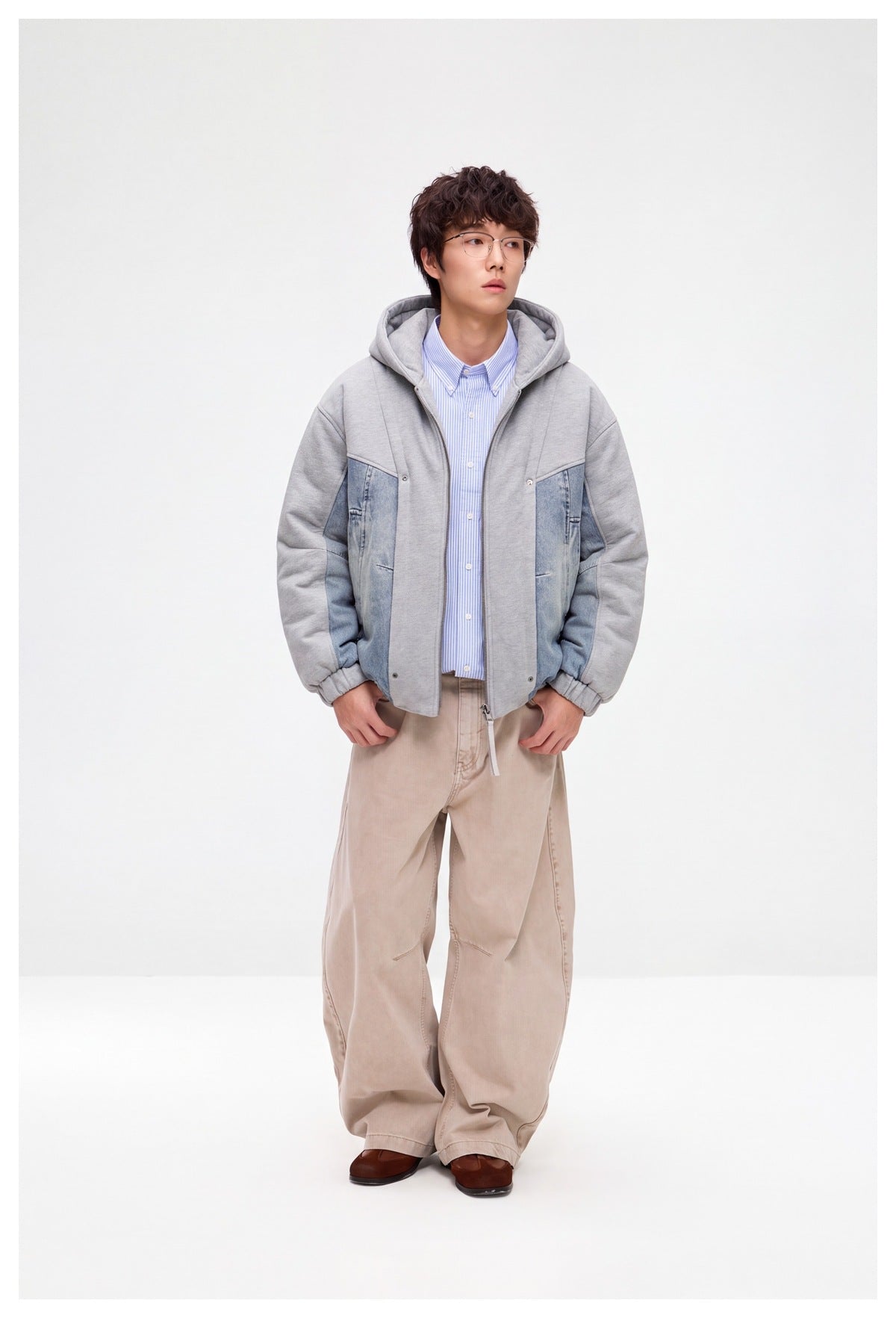 Full-length front view of a model wearing an open gray hooded jacket with blue denim panels and beige trousers.