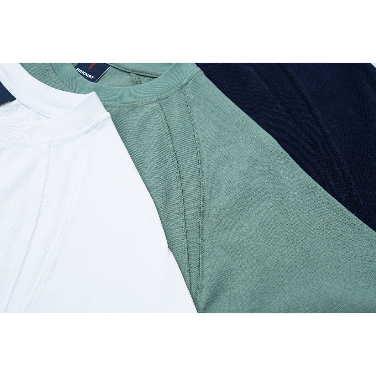 Close-up of white, sage green, and navy shirts showing their crew necks and raglan shoulder seams.