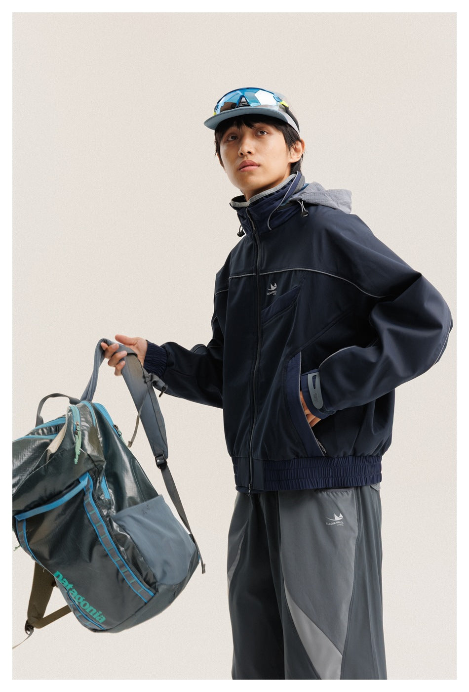 Model wearing a navy hooded jacket and gray trousers, holding a gray-blue backpack by its strap.