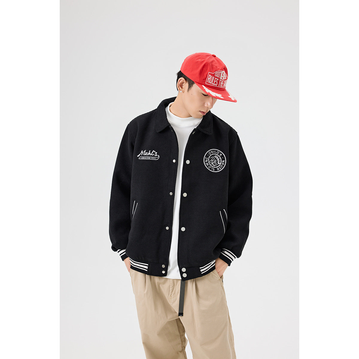 Model wearing a black varsity-style jacket with white striped trim, beige trousers and a red cap.