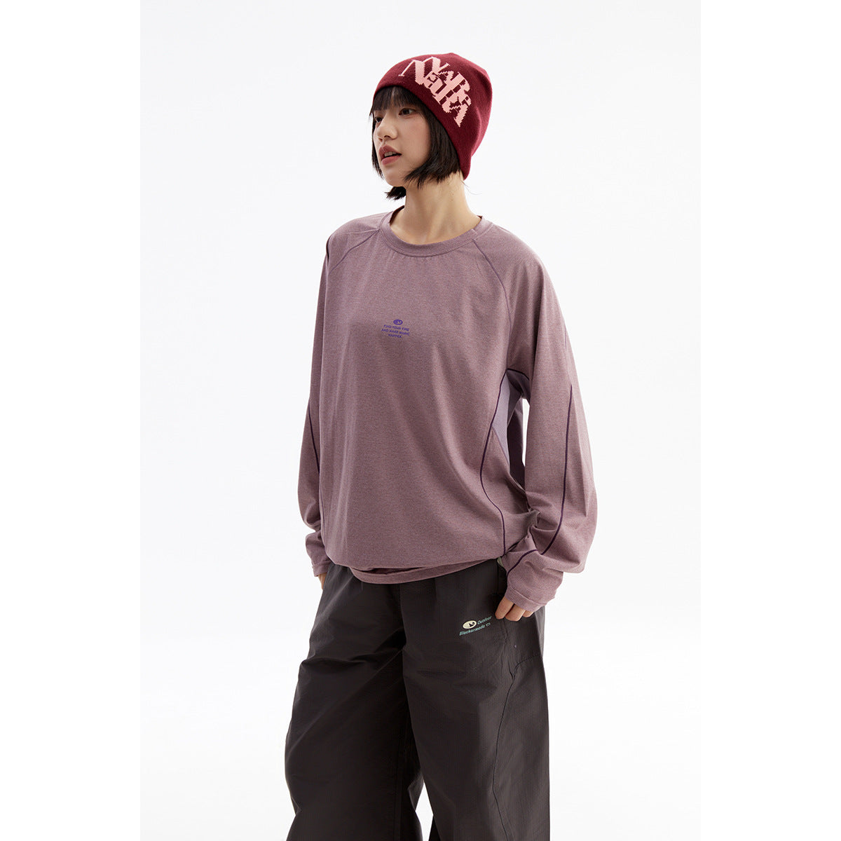 Model wearing a mauve long-sleeve crewneck top with dark piping, black pants, and a burgundy graphic beanie.