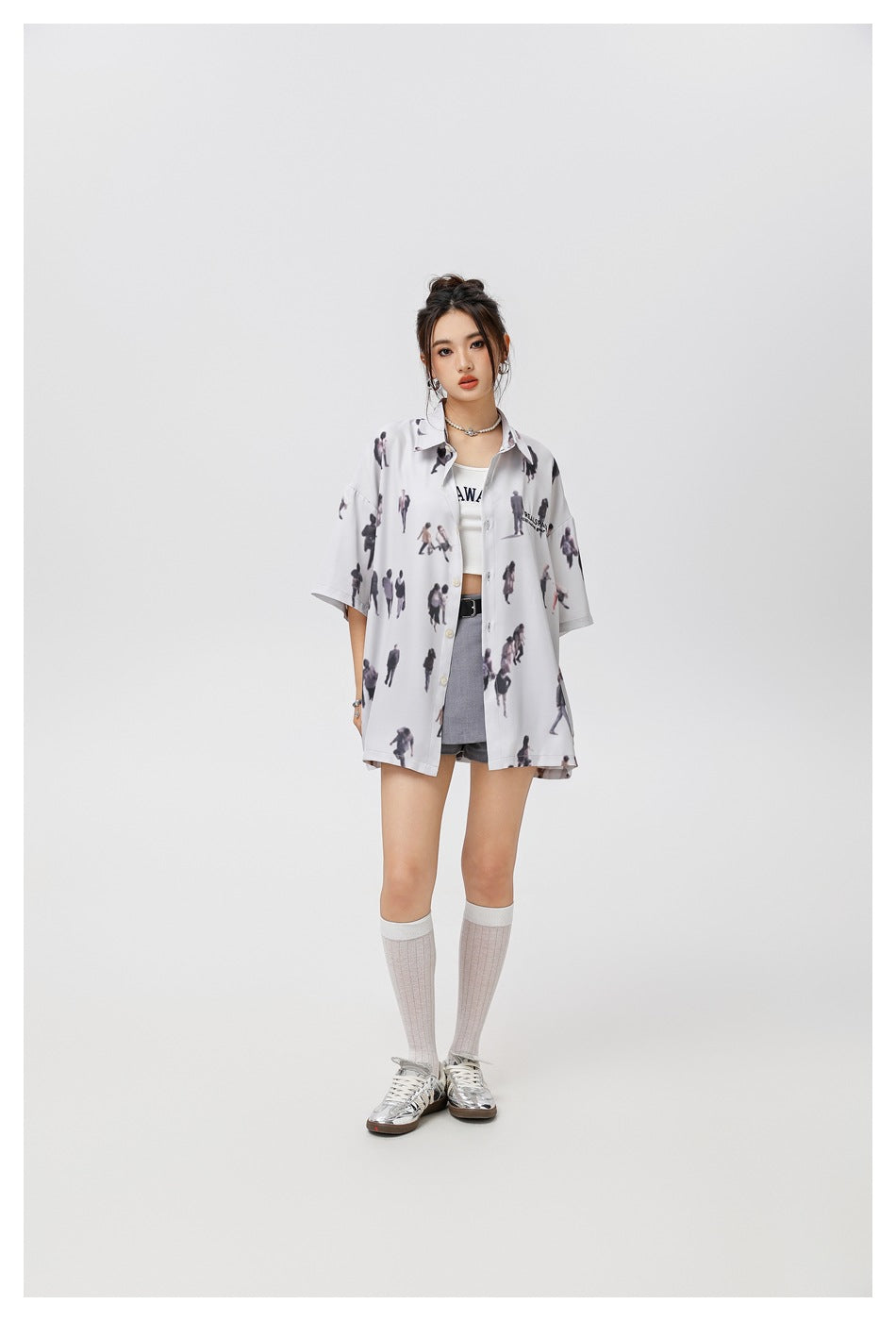Full-length model wearing a pale gray, miniature-people-print button-up shirt over a white top and gray shorts.