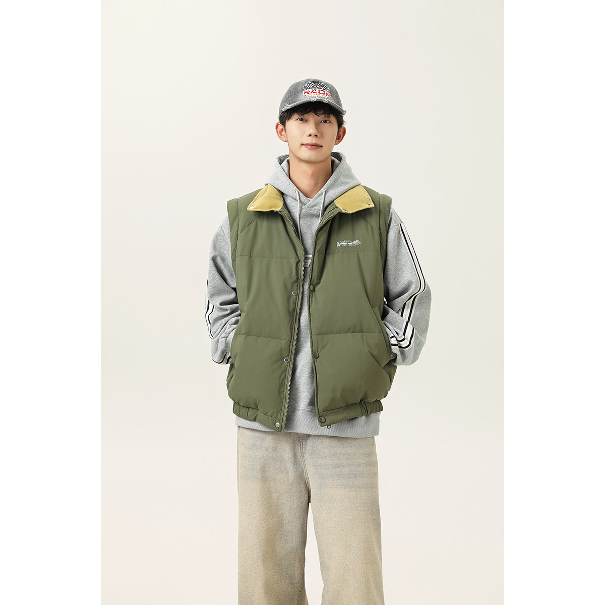 Model wearing an olive-green padded vest with a yellow-lined collar over a gray hoodie.