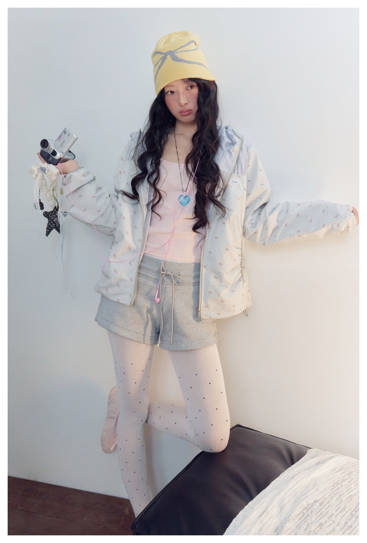 Model displays the front of a pale floral zip jacket with gray shorts and white dotted tights.