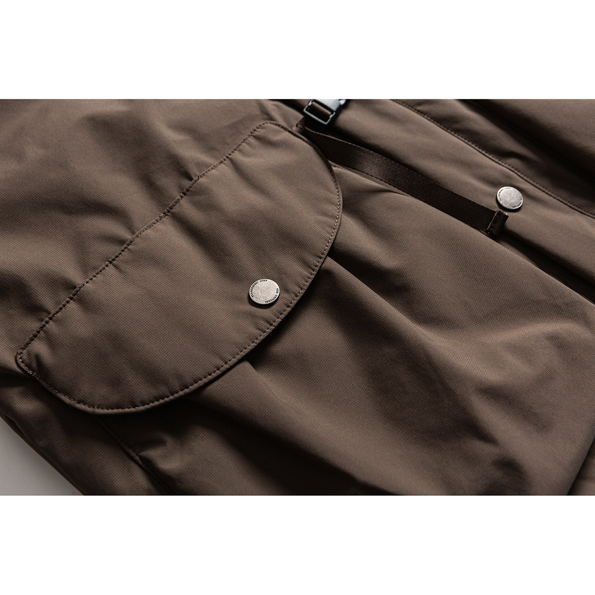 Close-up of a brown jacket’s curved flap pocket, metal snap buttons, and black adjustment strap.