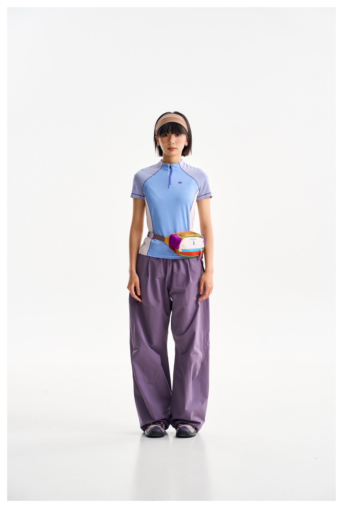 Model wearing a blue and lavender short-sleeve zip top, loose purple pants and a multicolored waist bag.