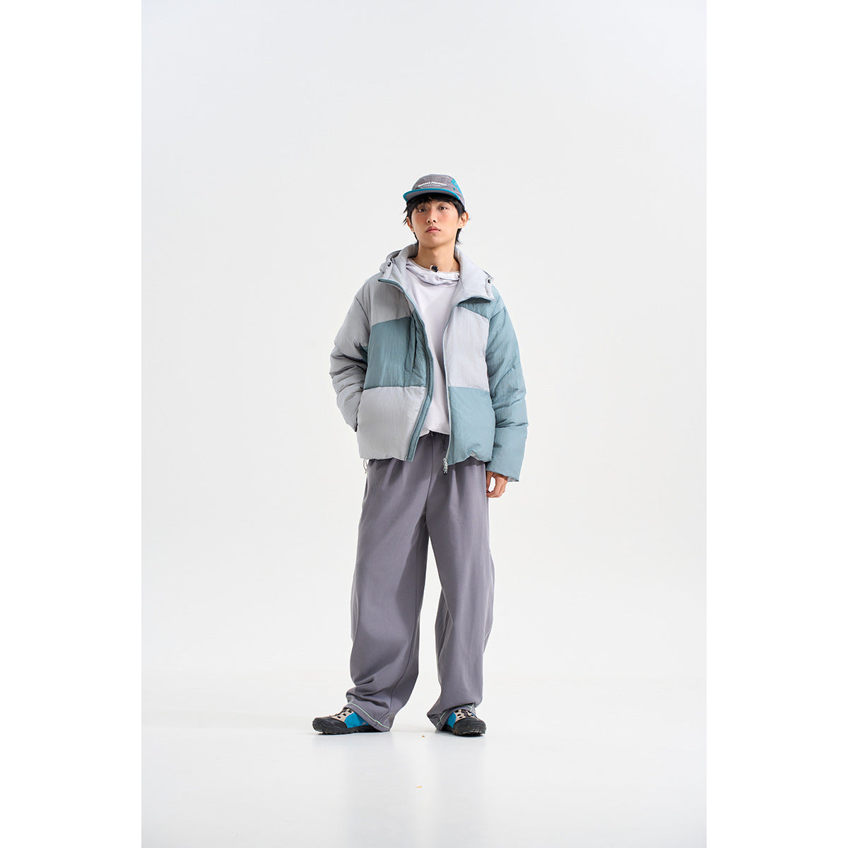 Model wearing a blue-gray color-block hooded puffer jacket over loose gray trousers.