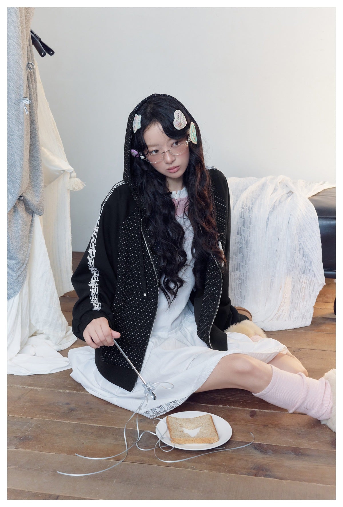 Model seated in a black hooded zip jacket with tiny white dots and white sleeve graphics, worn over a white dress.