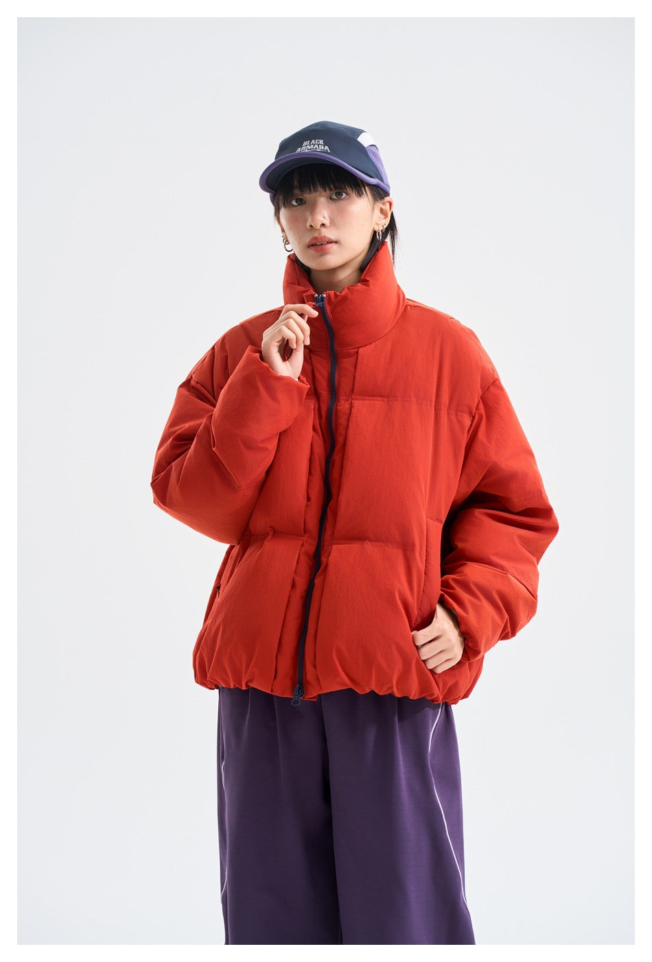 Model wearing a red-orange puffer jacket with a high collar and purple trousers, shown from the front.