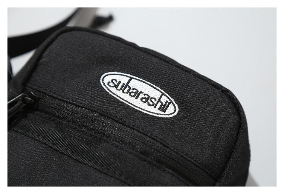 Close-up of a black bag with an oval embroidered label and a horizontal zip pocket.