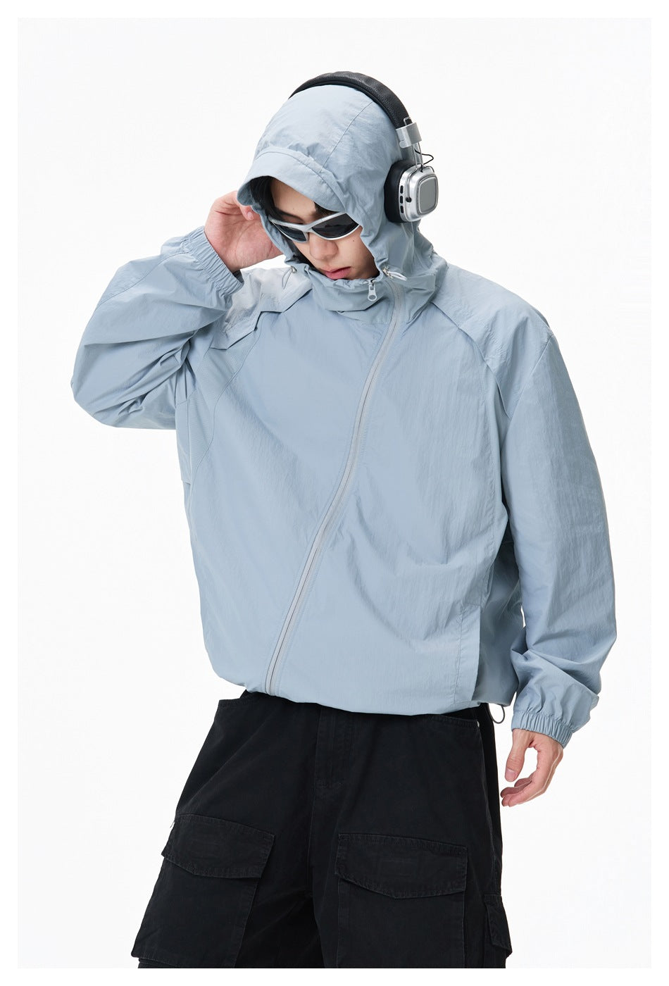 Model wearing a pale blue-gray hooded jacket with an angled zipper and elastic cuffs, paired with black cargo pants.