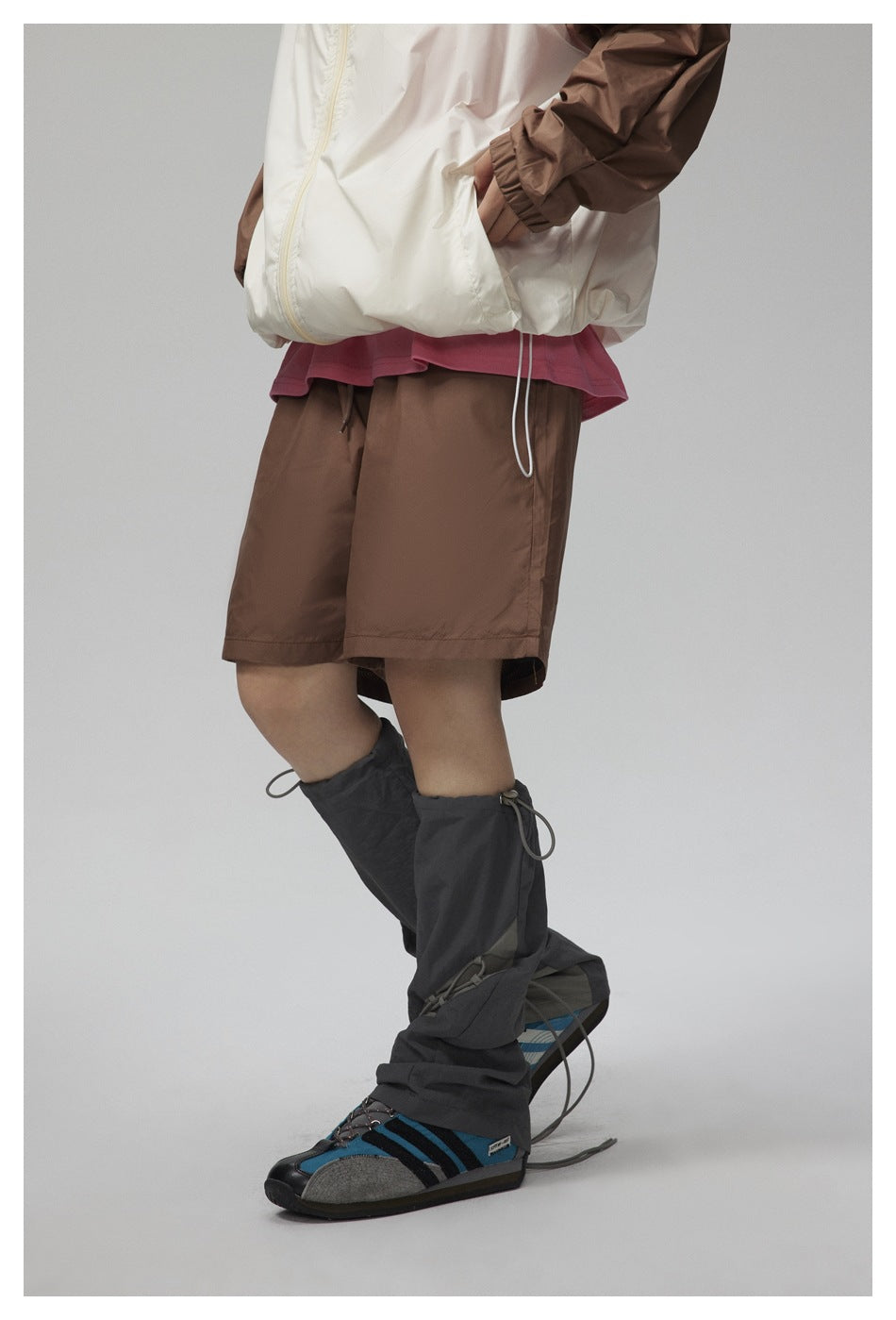 Cropped front three-quarter view of a white jacket with brown sleeves, brown shorts, and dark gaiters over blue shoes.