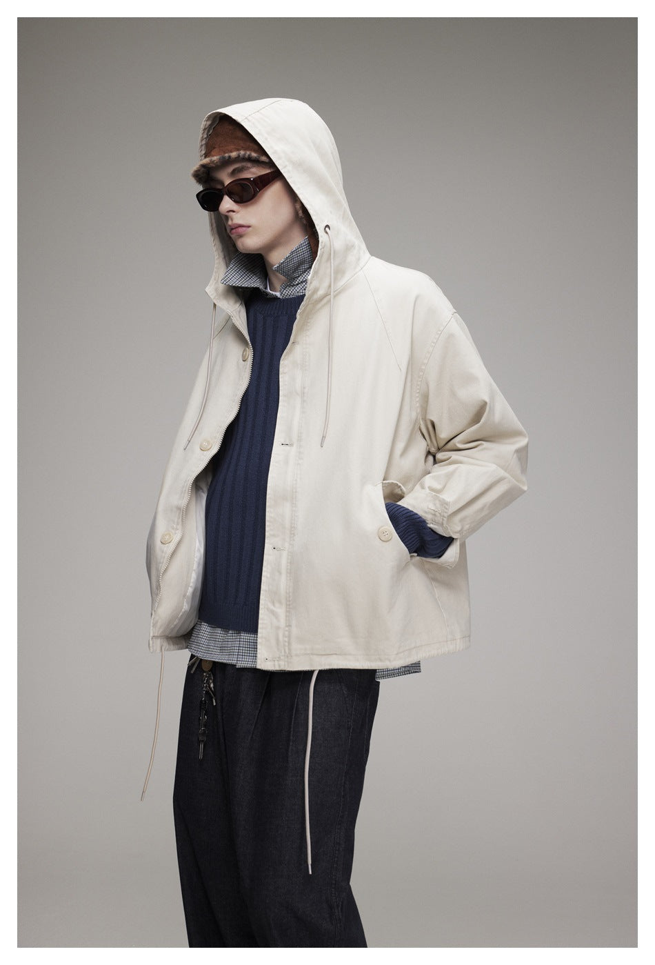 Model wearing an off-white hooded jacket over a navy sweater and checked shirt, with dark trousers.