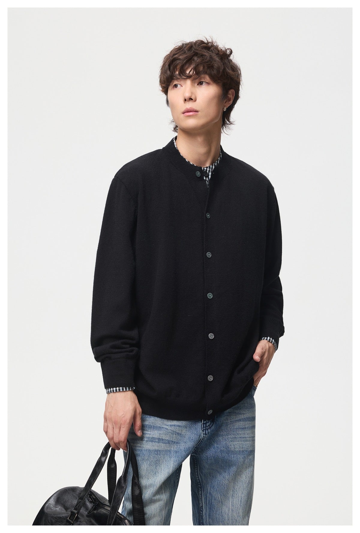 Man wearing black button-front cardigan over a checked collar layer with blue jeans, holding a black bag