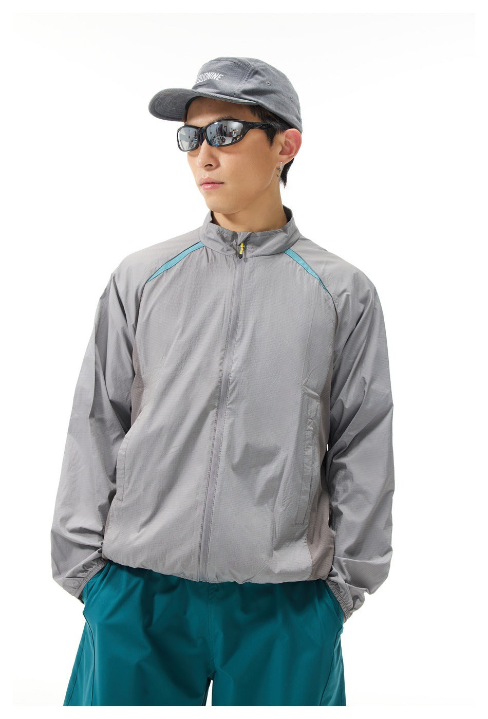 Man wearing a light gray zip-up jacket with turquoise shoulder accents and teal trousers, shown from the front.