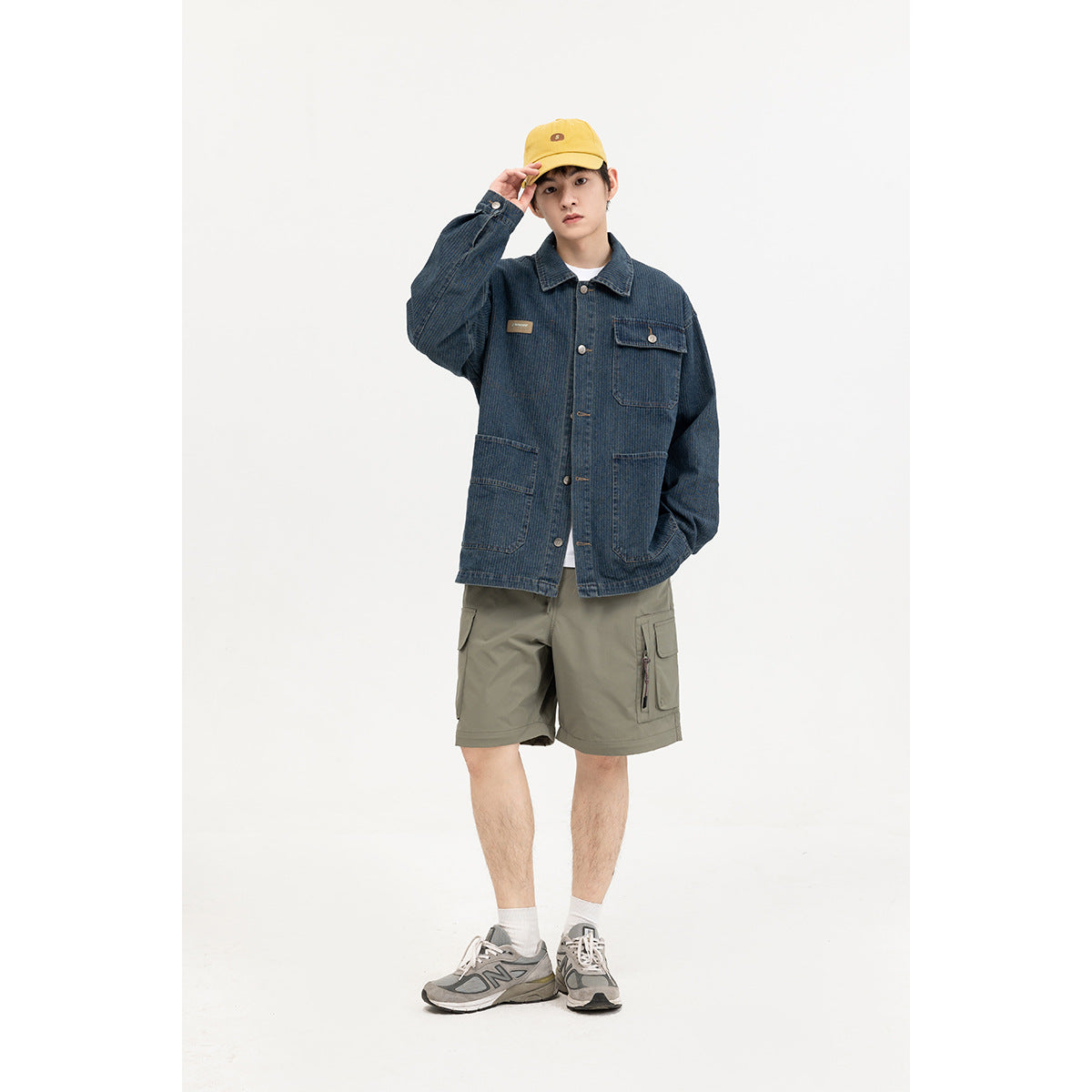 Model wearing a dark blue denim chore jacket with patch pockets, olive cargo shorts, and a yellow cap.