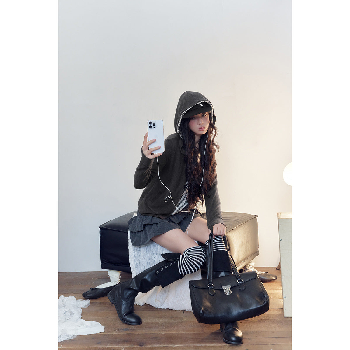 Model seated in a charcoal hooded zip jacket with striped lining, striped top, tiered dark skirt, and striped knee socks.