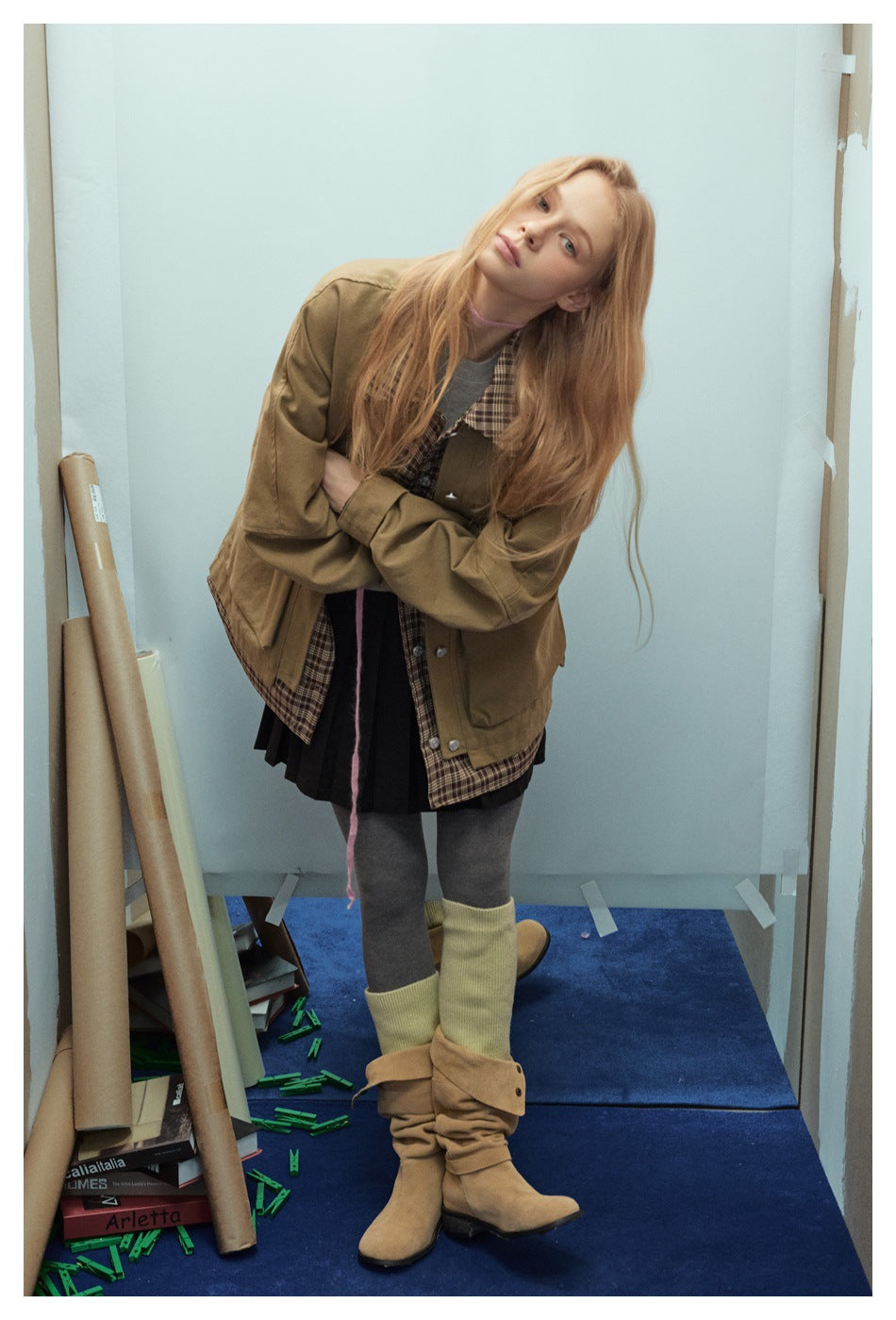 Model wearing a tan oversized jacket with plaid collar and hem, a black pleated skirt, gray tights, and tan boots.