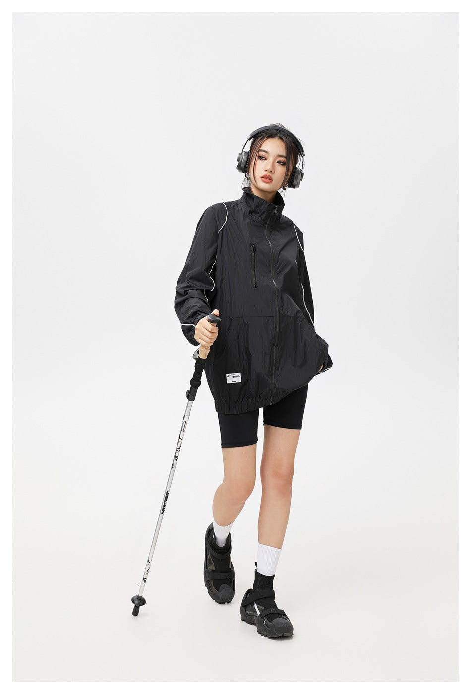 Model wearing a black jacket with white piping, posed with a trekking pole and headphones.