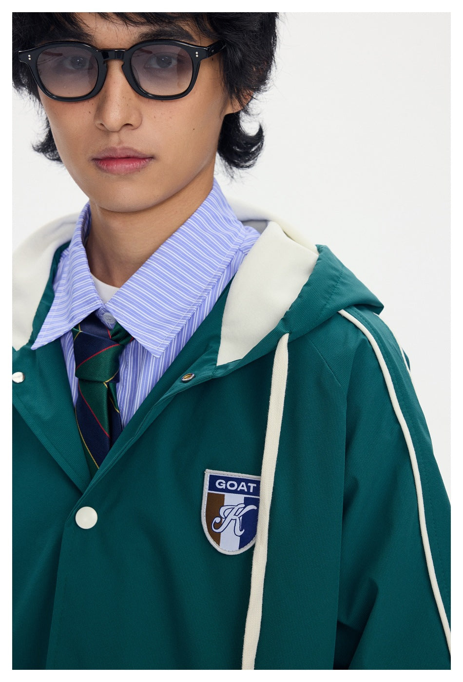 Close-up of a model wearing a teal hooded jacket with cream piping and a GOAT chest patch, over a striped shirt and tie.