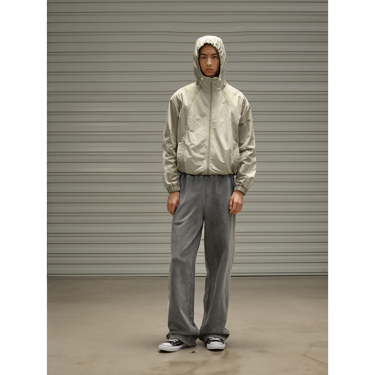 Model wearing a light beige hooded zip jacket and loose charcoal gray pants, standing front-facing.