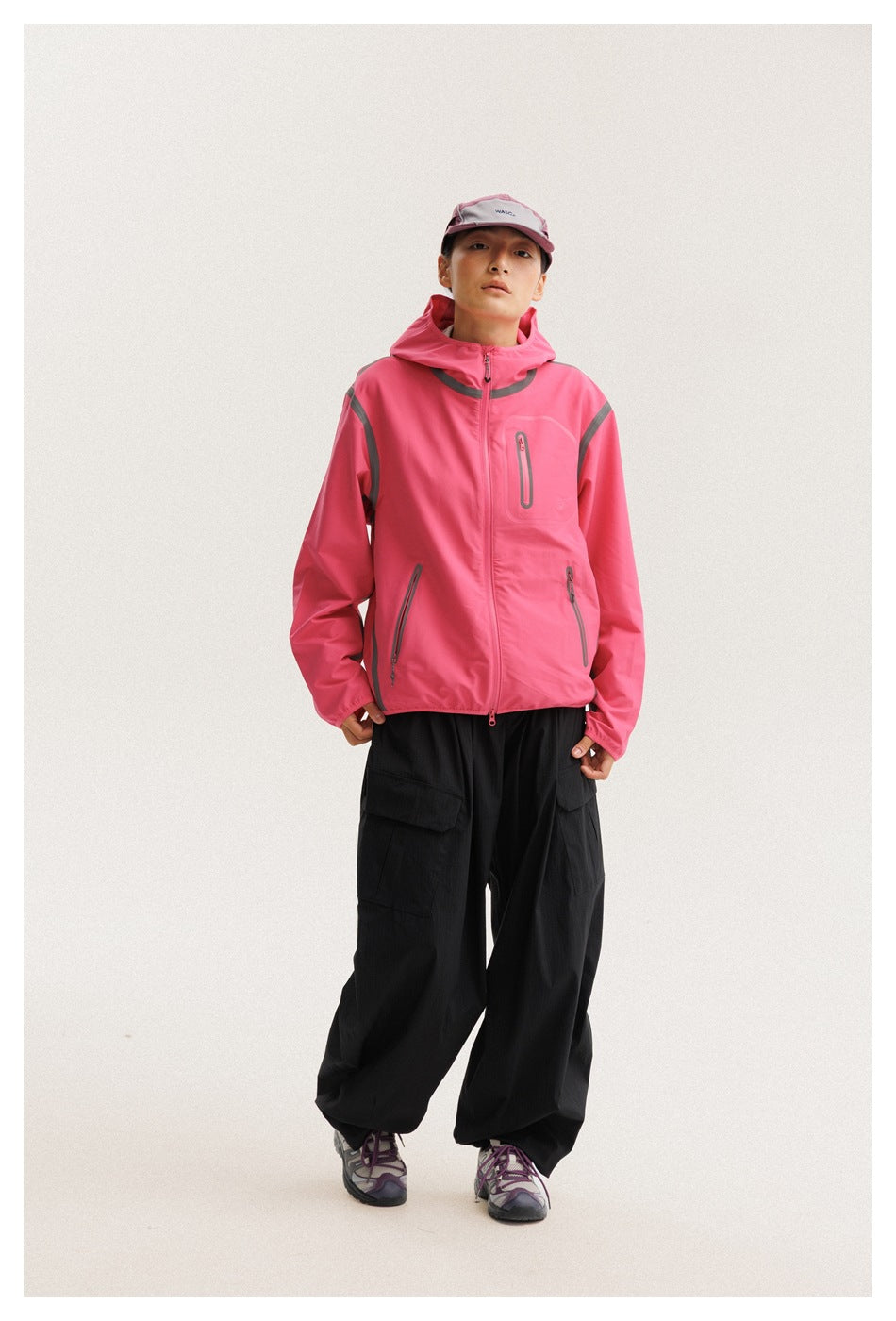 Front view of a model wearing a bright pink hooded jacket with gray trim and loose black cargo trousers.