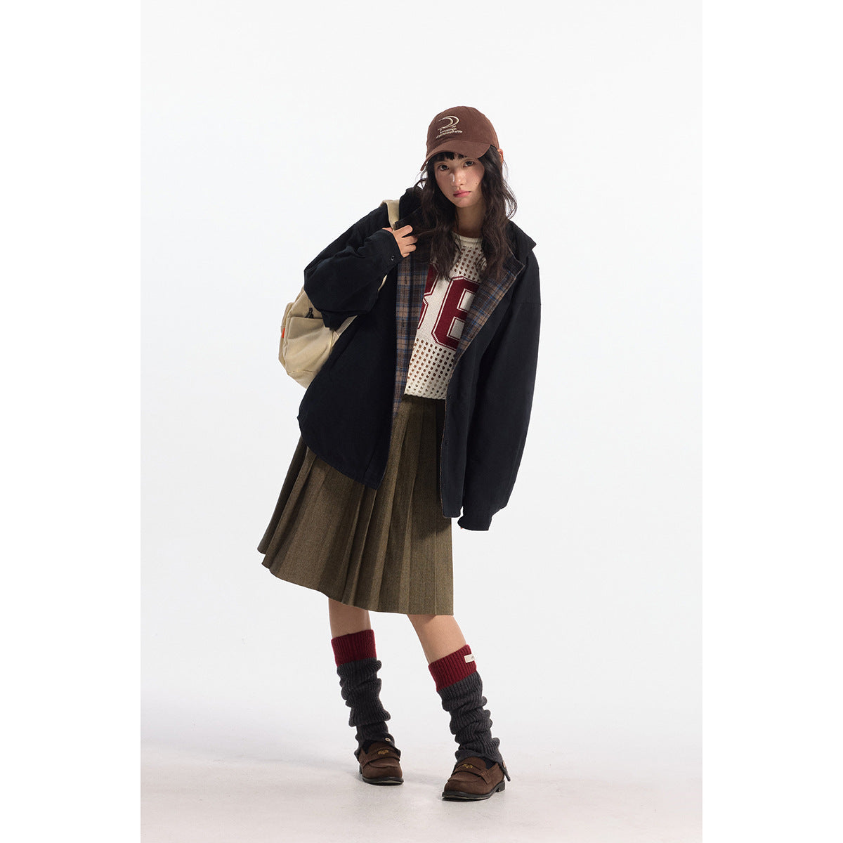 Model wearing a loose black jacket with plaid lining, a cream mesh top, and an olive pleated skirt.