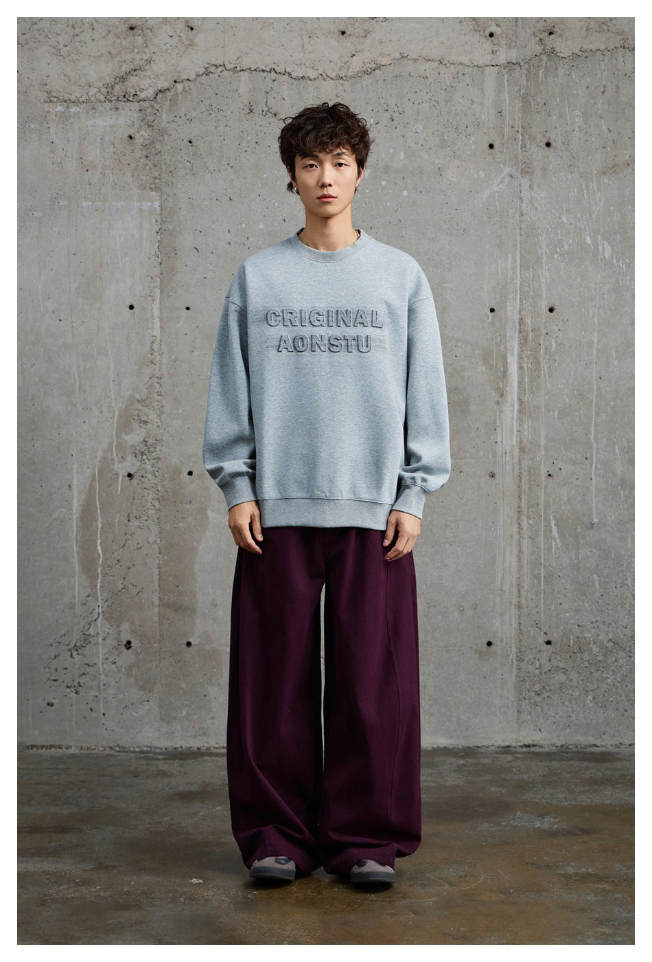Front view of a model wearing a light gray crewneck sweatshirt with raised lettering and loose burgundy trousers.