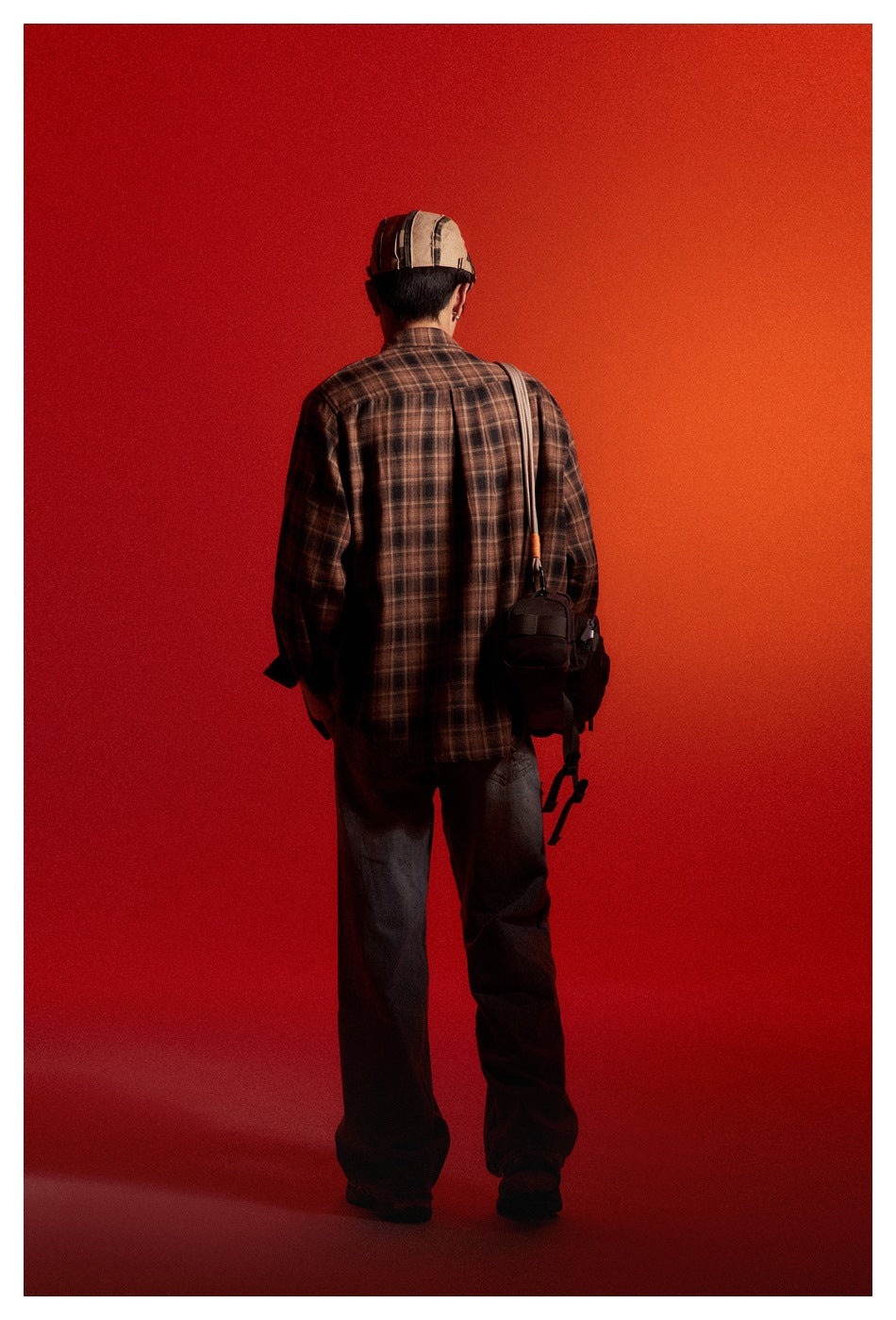 Back view of a model wearing a dark brown plaid shirt, loose dark jeans and a striped cap, with a shoulder bag.
