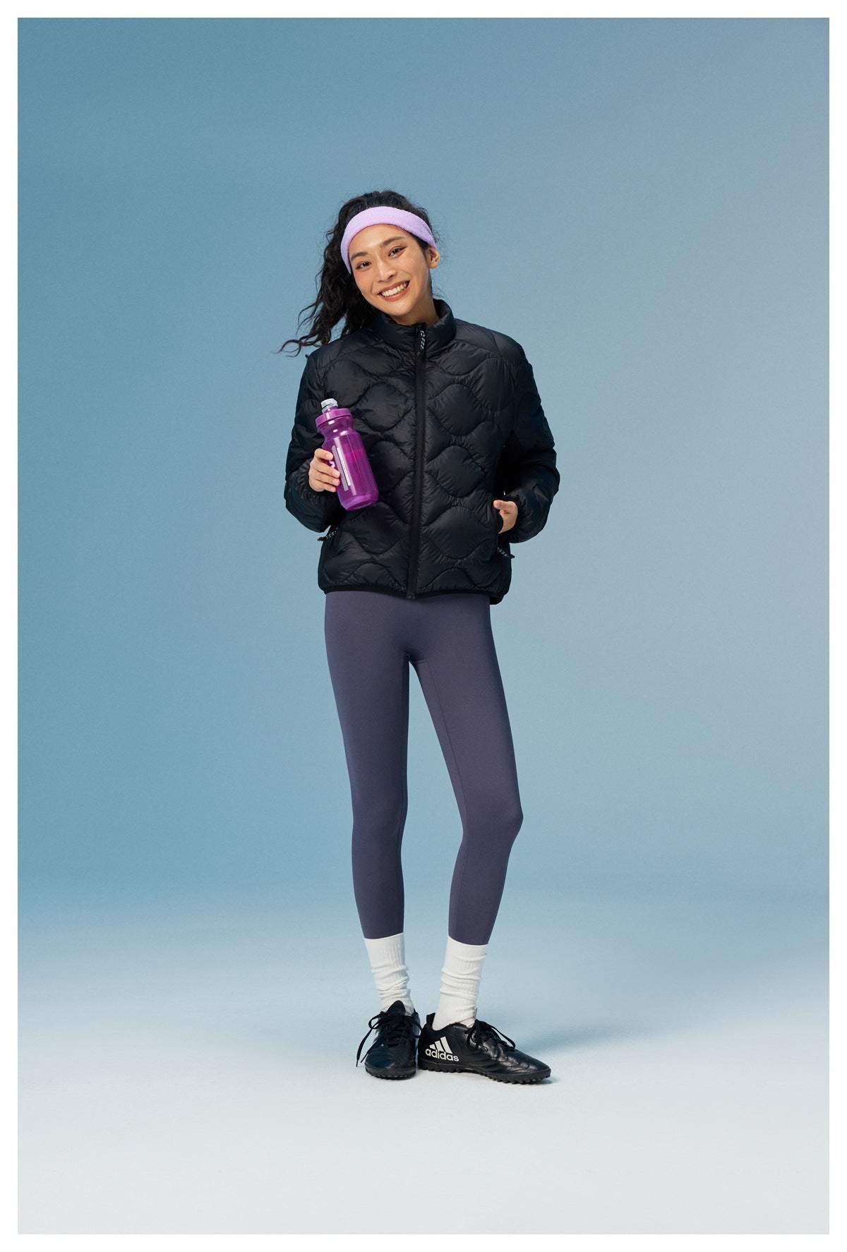Woman wearing the black quilted zip jacket with leggings and a purple headband, holding a water bottle