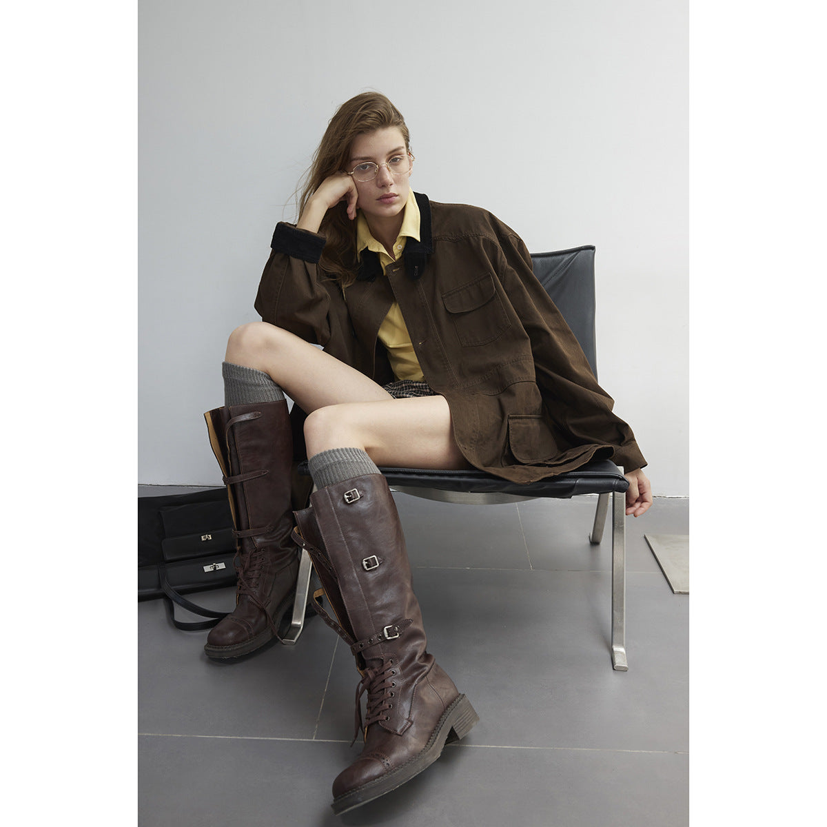 Model seated in an oversized dark brown utility jacket with a black collar and flap pockets.