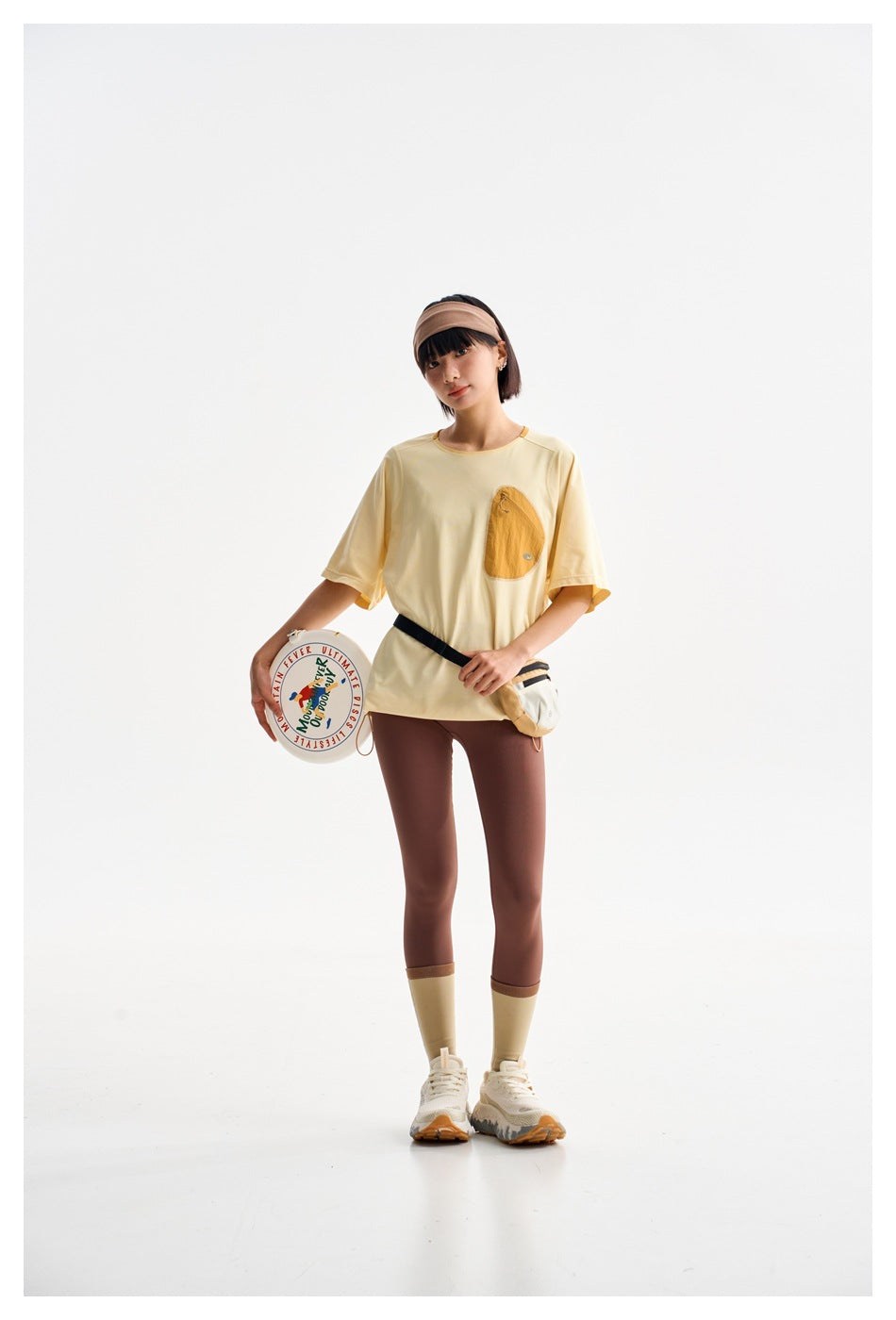 Model wearing a pale yellow short-sleeve tee with a chest pocket and brown leggings, holding a printed disc.