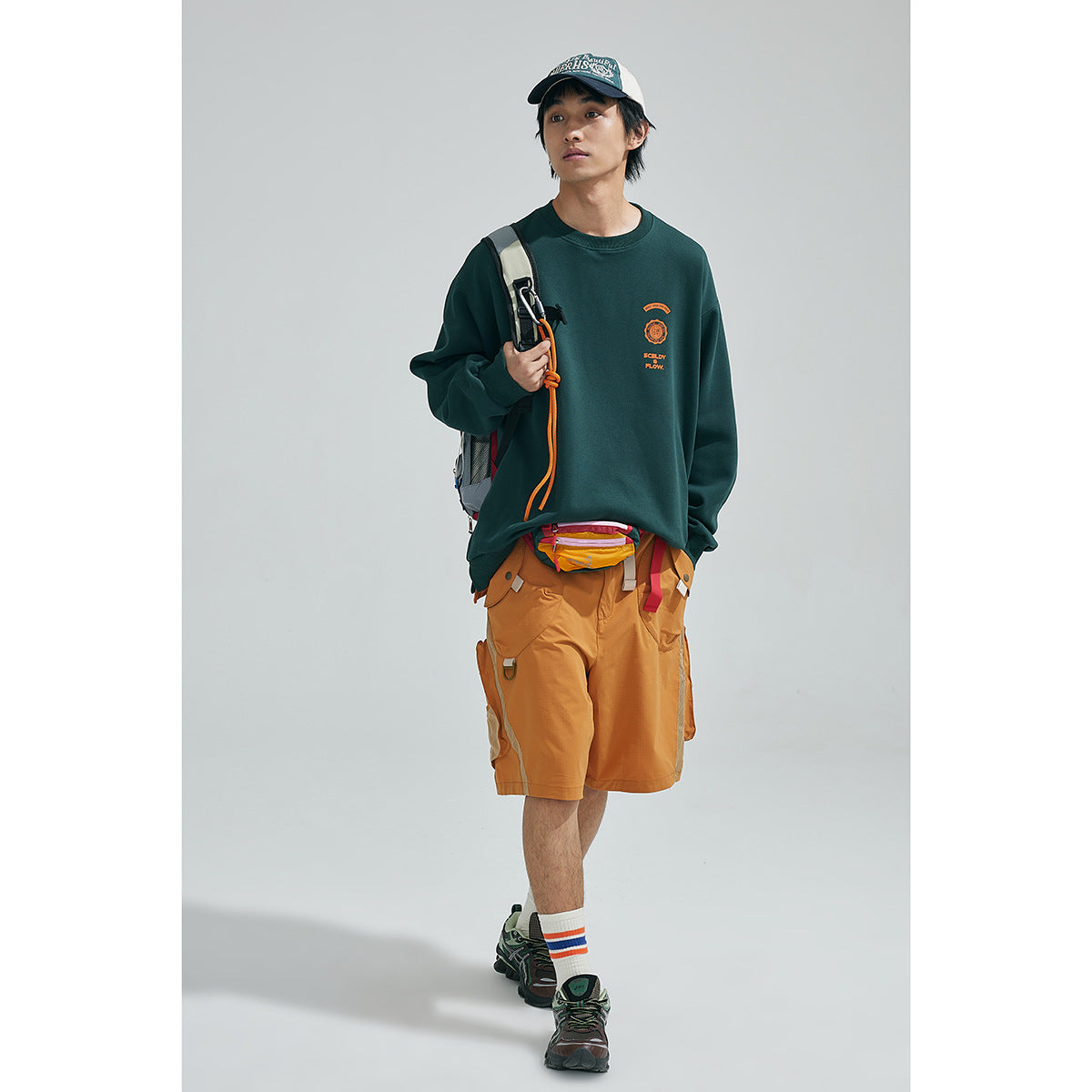 Model wearing a dark green sweatshirt with orange chest print and ochre cargo shorts, styled with a cap and backpack.