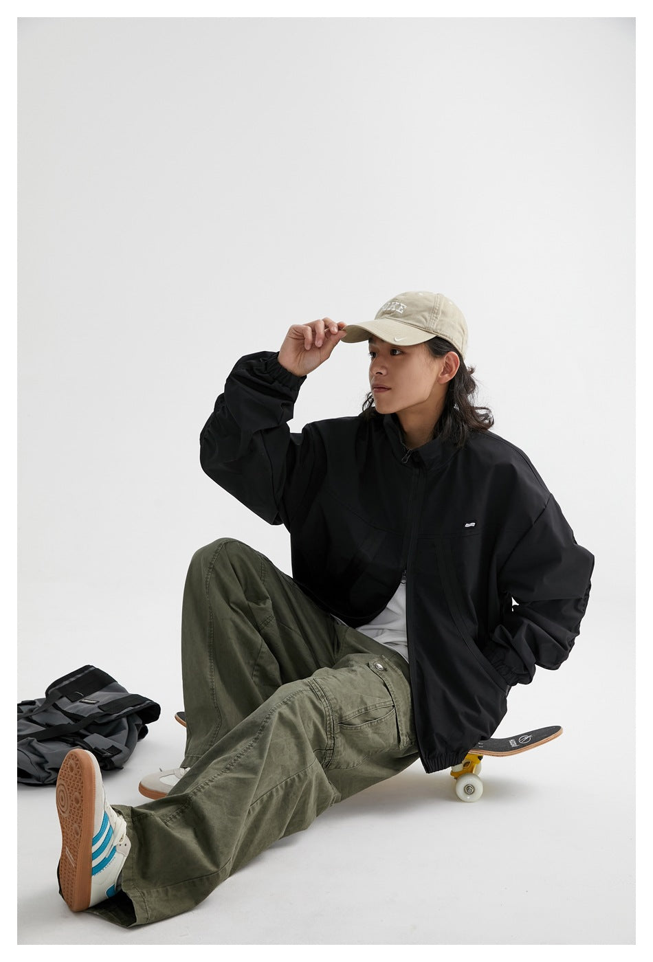 Seated model wearing a black zip-front jacket with elastic cuffs and hem, with a backpack and skateboard nearby.