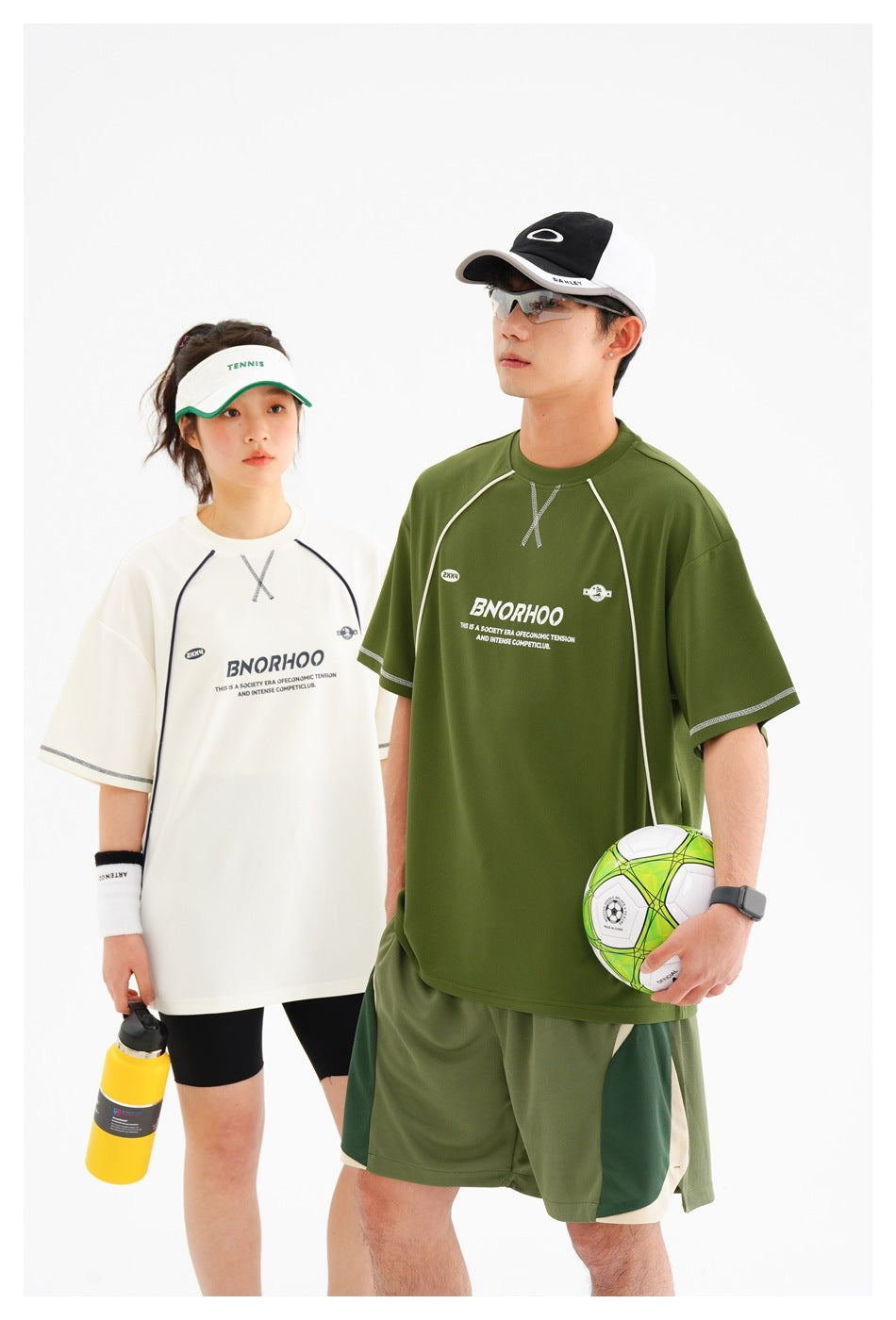 Two models pose in white and olive-green graphic sports T-shirts; the man also wears matching shorts.