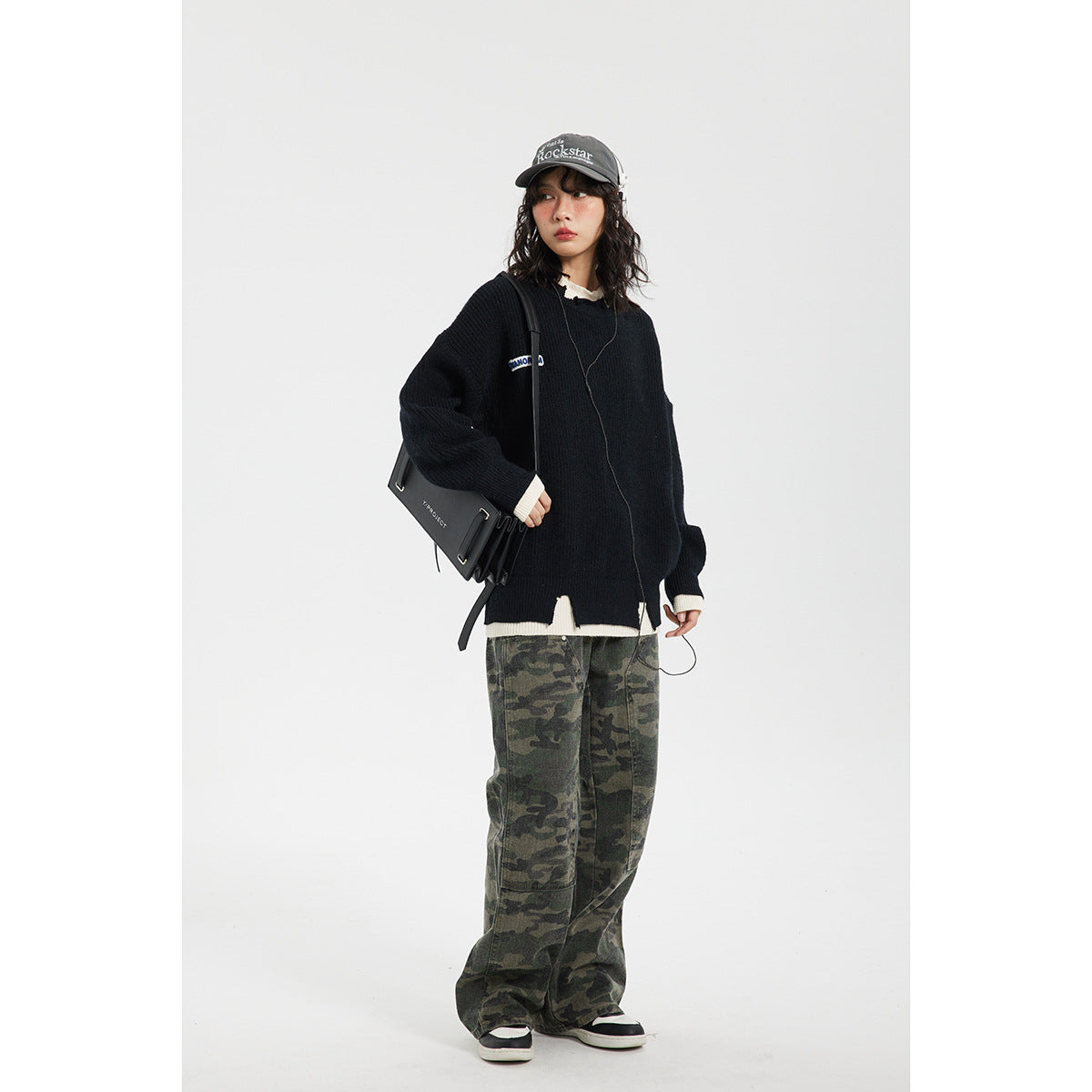 Model wearing an oversized black sweater with cream trim and loose green camouflage trousers.