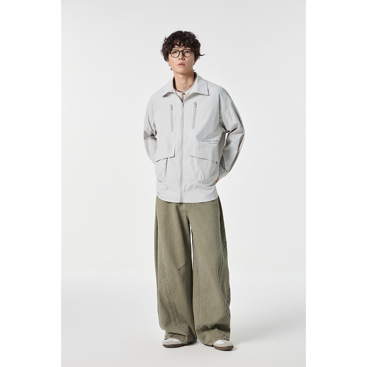 Full-length model wearing a light gray utility jacket with zip pockets and loose olive trousers.