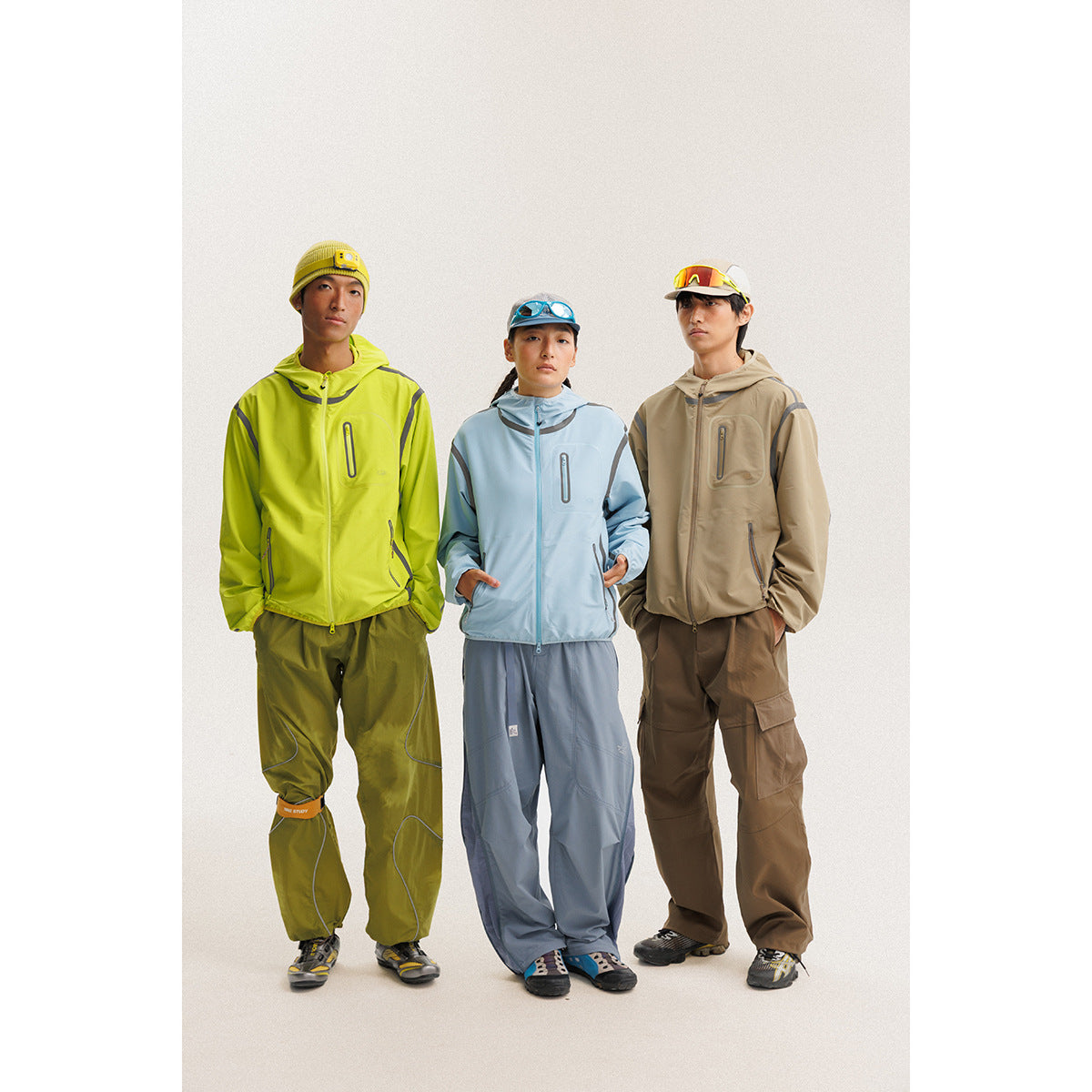 Three models wear hooded jackets and loose outdoor pants in lime green, pale blue, and tan.