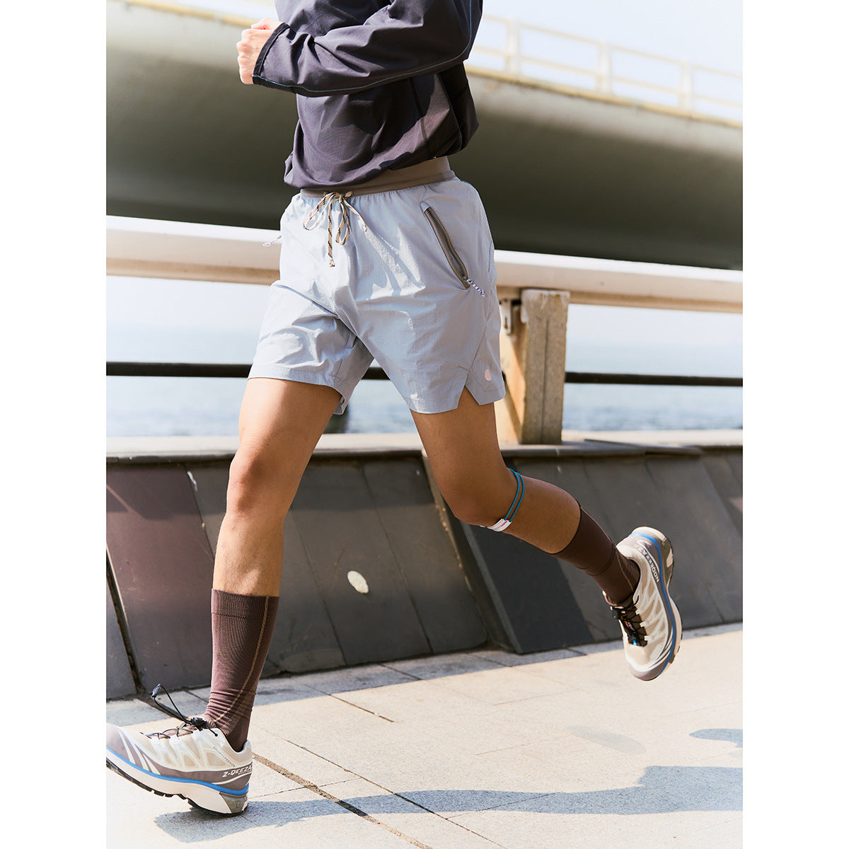 Person running in light gray drawstring shorts with diagonal zip pockets, shown from waist to shoes.