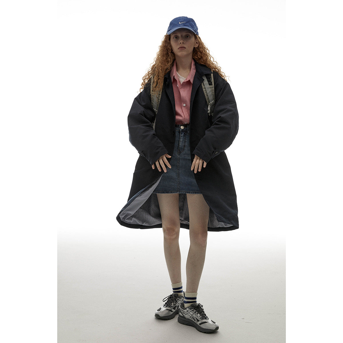 Model wearing a long black coat over a pink shirt and denim skirt, with a blue cap and backpack.