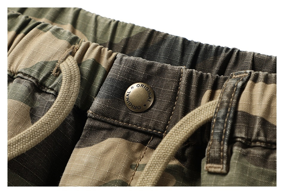 Close-up of the camouflage shorts' gathered waistband, drawstring, belt loop, and metal button engraved “ORIGINAL.”
