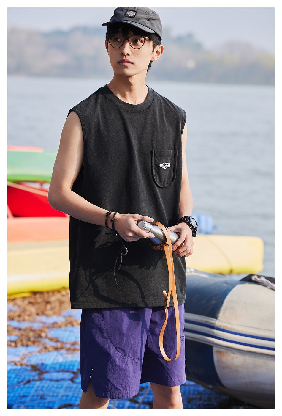 Front view of a model wearing a black sleeveless pocket shirt and purple shorts by the water.