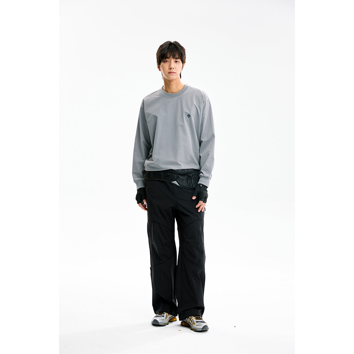 Model wearing a gray long-sleeve crewneck with a small chest emblem and loose black paneled pants.