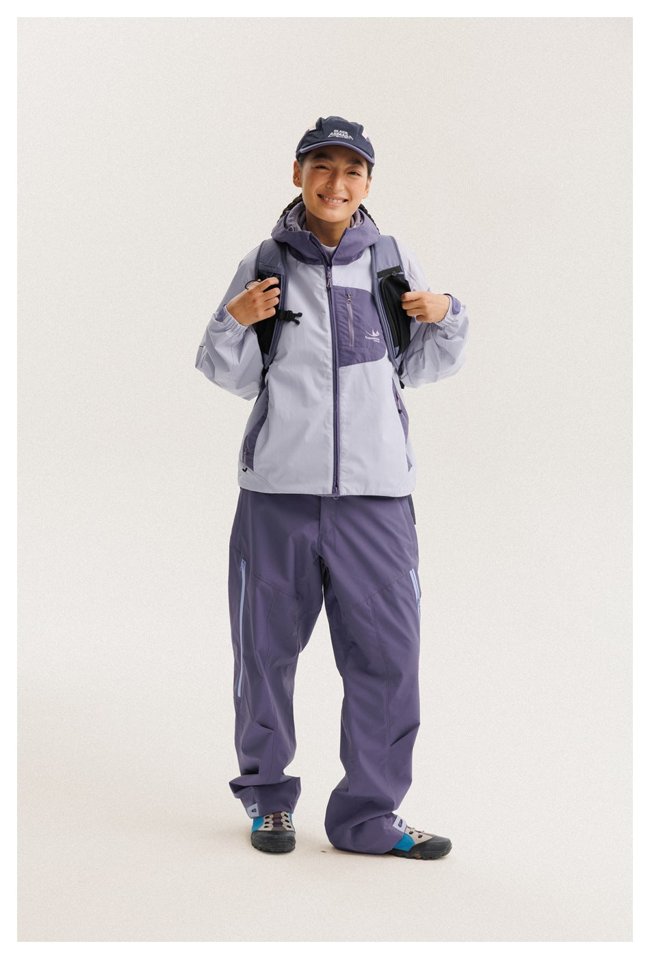 Front view of a model wearing a light gray and purple hooded jacket with matching purple trousers and a backpack.