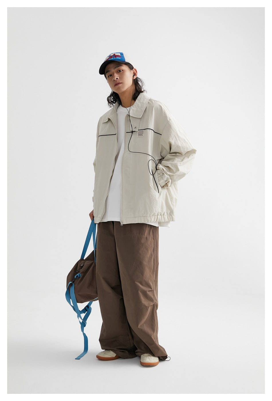 Full-length model wearing an ivory piped jacket and brown trousers, carrying a brown bag with blue straps.