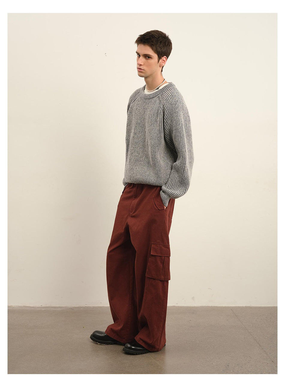 Male model wearing a gray ribbed crewneck sweater and loose rust-red cargo pants, posed in profile.