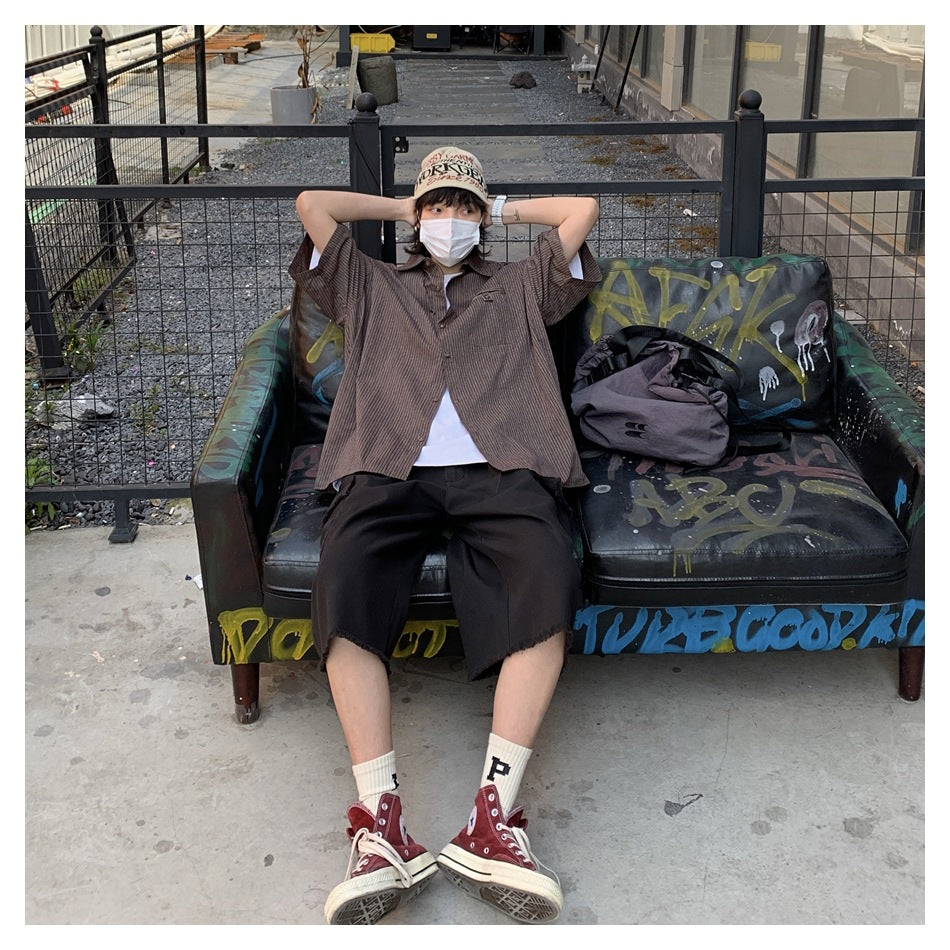 Masked model seated outdoors in a loose dark brown striped shirt, black shorts, and red high-top sneakers.