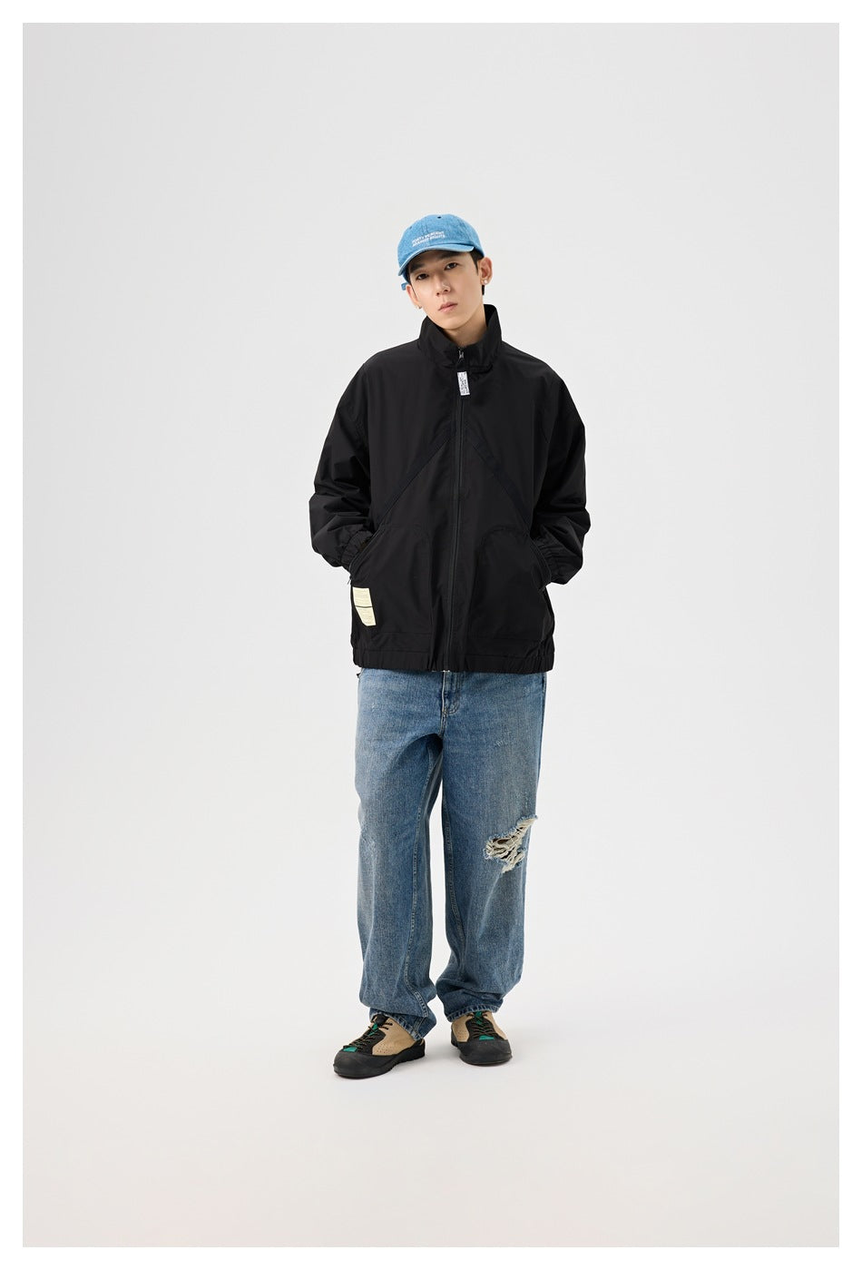 Full-length front view of a model wearing a black high-collar zip jacket, loose blue jeans, and a blue cap.